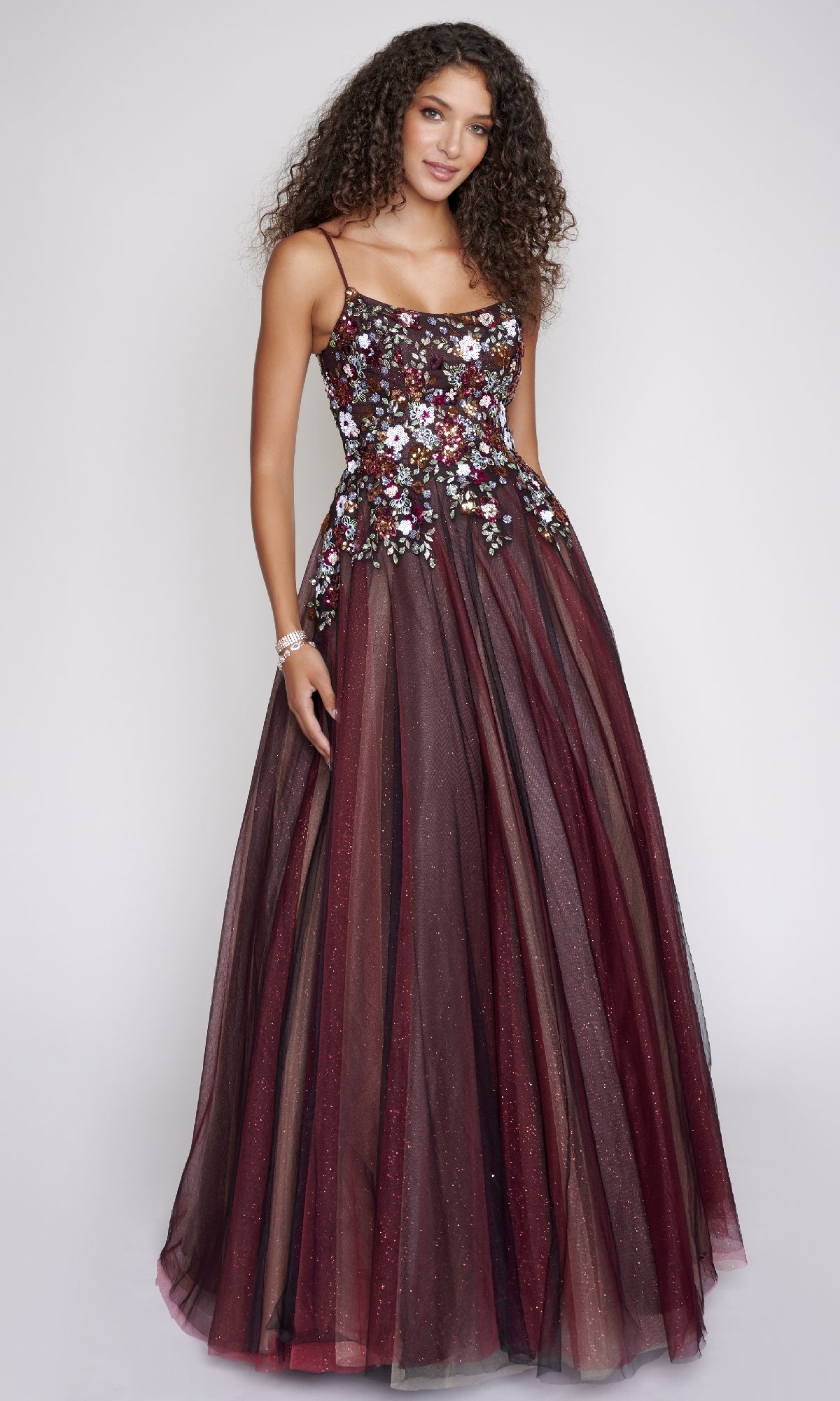 A woman in the Nina Canacci 3206 prom ball gown—burgundy with thin straps, a floral embroidered bodice, and a glitter tulle skirt—stands against a plain light background.